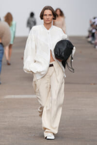 Model on a runway wearing a white oversized quilted jacket and beige wide trousers, holding a black chain-strap bag.