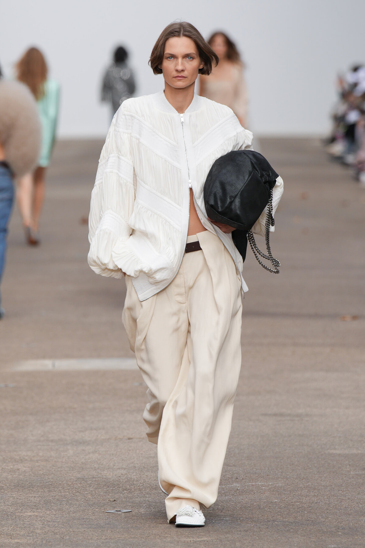 Model on a runway wearing a white oversized quilted jacket and beige wide trousers, holding a black chain-strap bag.