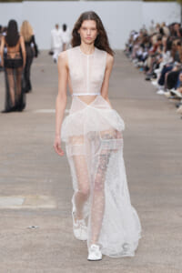 Model walking a runway in a sheer white lace gown with a sheer overlay and floral details, audience seated along the sides.
