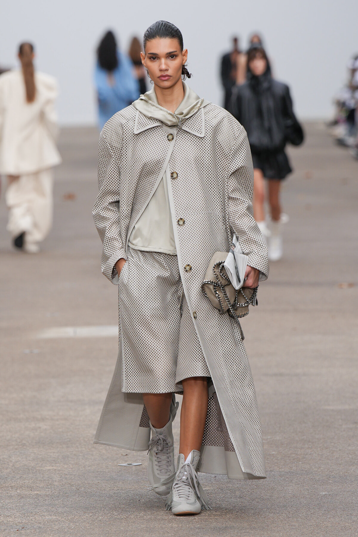 Model walks a runway in a beige perforated coat with matching wide-leg trousers, light scarf, and gray high-top sneakers.