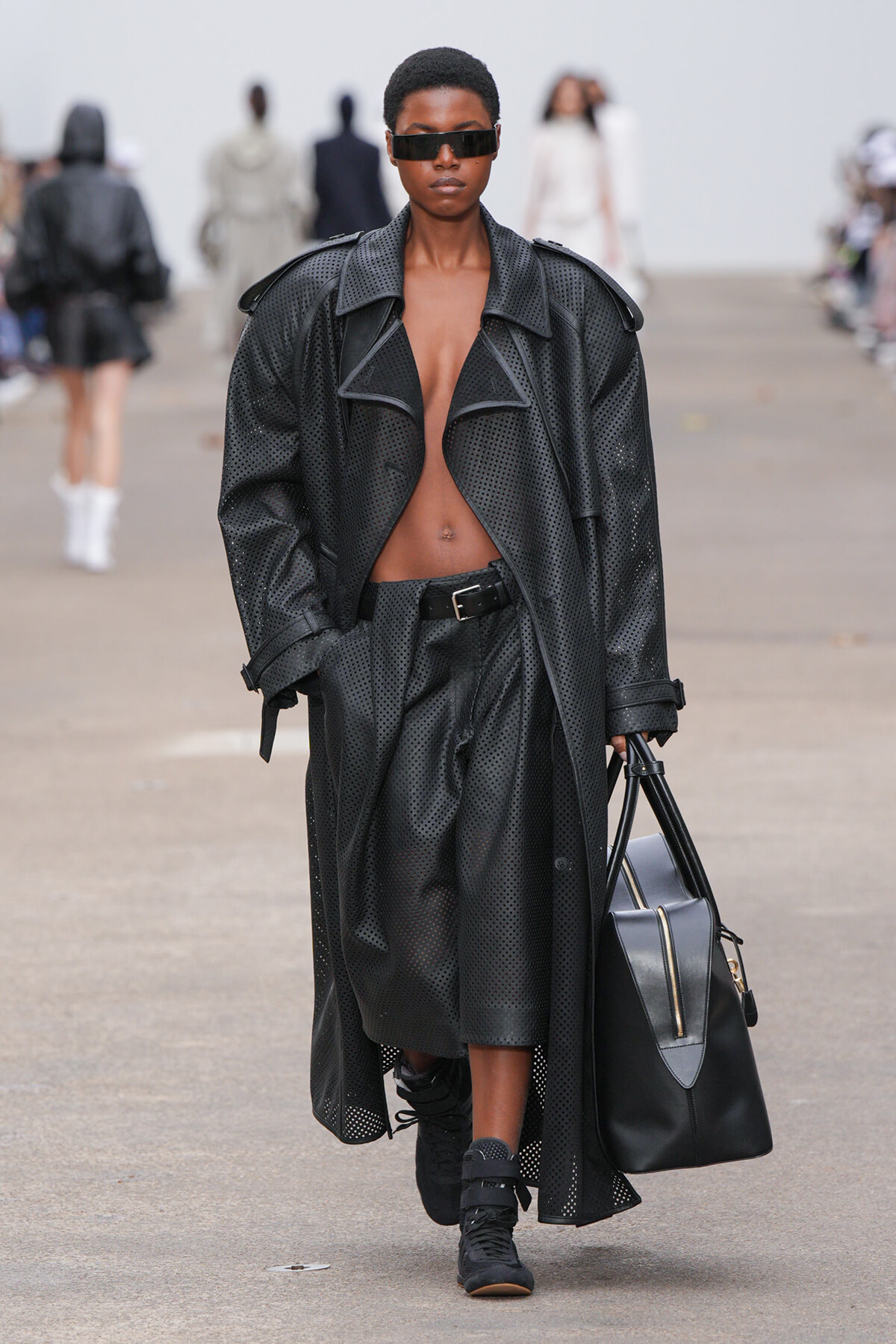 Model walks a runway in a black perforated trench coat, sunglasses, and a large black handbag.