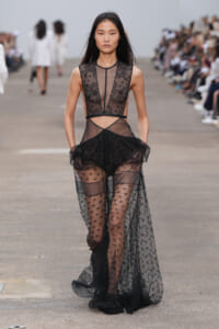 Model walking a runway in a sheer black lace gown with cutouts and a flowing train on a fashion show catwalk, focused expression.