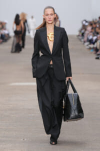 Model walking a runway in a black suit with plunging shirt-front, carrying a black structured handbag; oversized gold statement necklace visible.