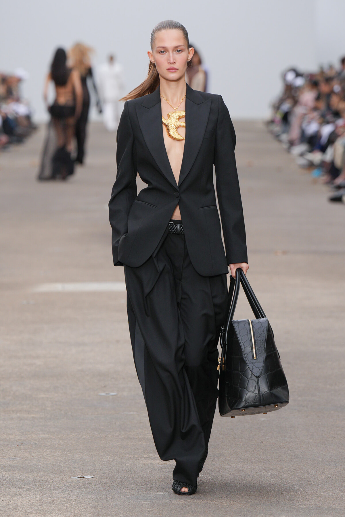 Model walking a runway in a black suit with plunging shirt-front, carrying a black structured handbag; oversized gold statement necklace visible.
