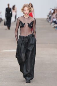 Model on a runway wearing a sheer black mesh top with cup-like chest panels and oversized perforated black trousers, hands in pockets.