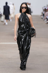 Model walks a fashion show in a glossy black halter jumpsuit with oversized sunglasses, holding a black clutch.