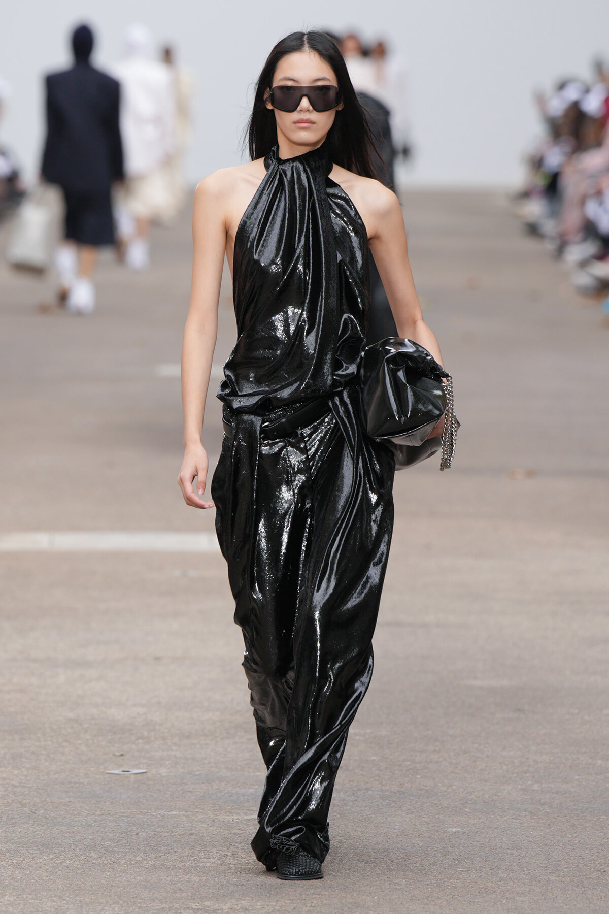 Model walks a fashion show in a glossy black halter jumpsuit with oversized sunglasses, holding a black clutch.