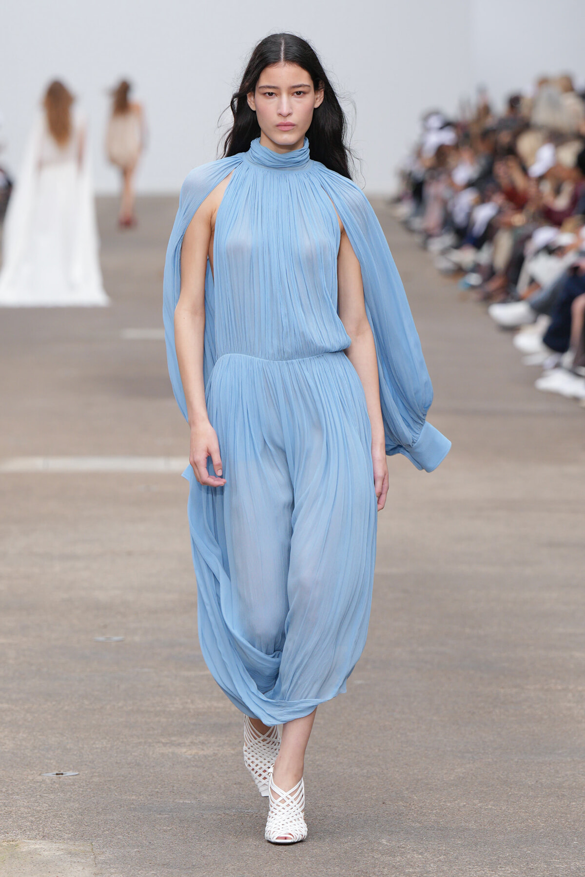 Model walks the runway in a light blue, pleated draped jumpsuit with cape-like sleeves and high neckline.