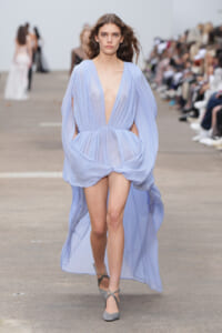Model walks the runway in a pale blue, plunging V-neck gown with draped, billowing sleeves and a short front overlong, flowing train.