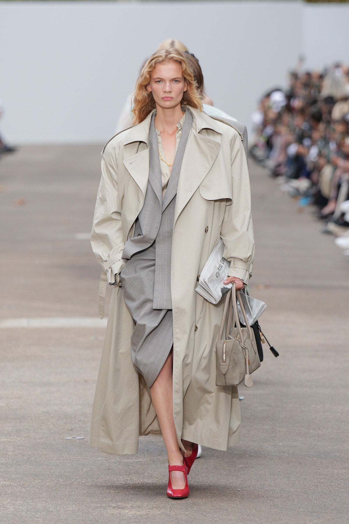 Model walking a fashion show in a beige trench over a gray suit, carrying a newspaper and light-gray bag with red shoes.