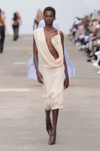 Model walking a runway in a sleeveless, draped beige dress with a gold strap and deep относительно neckline, wearing gray heels.