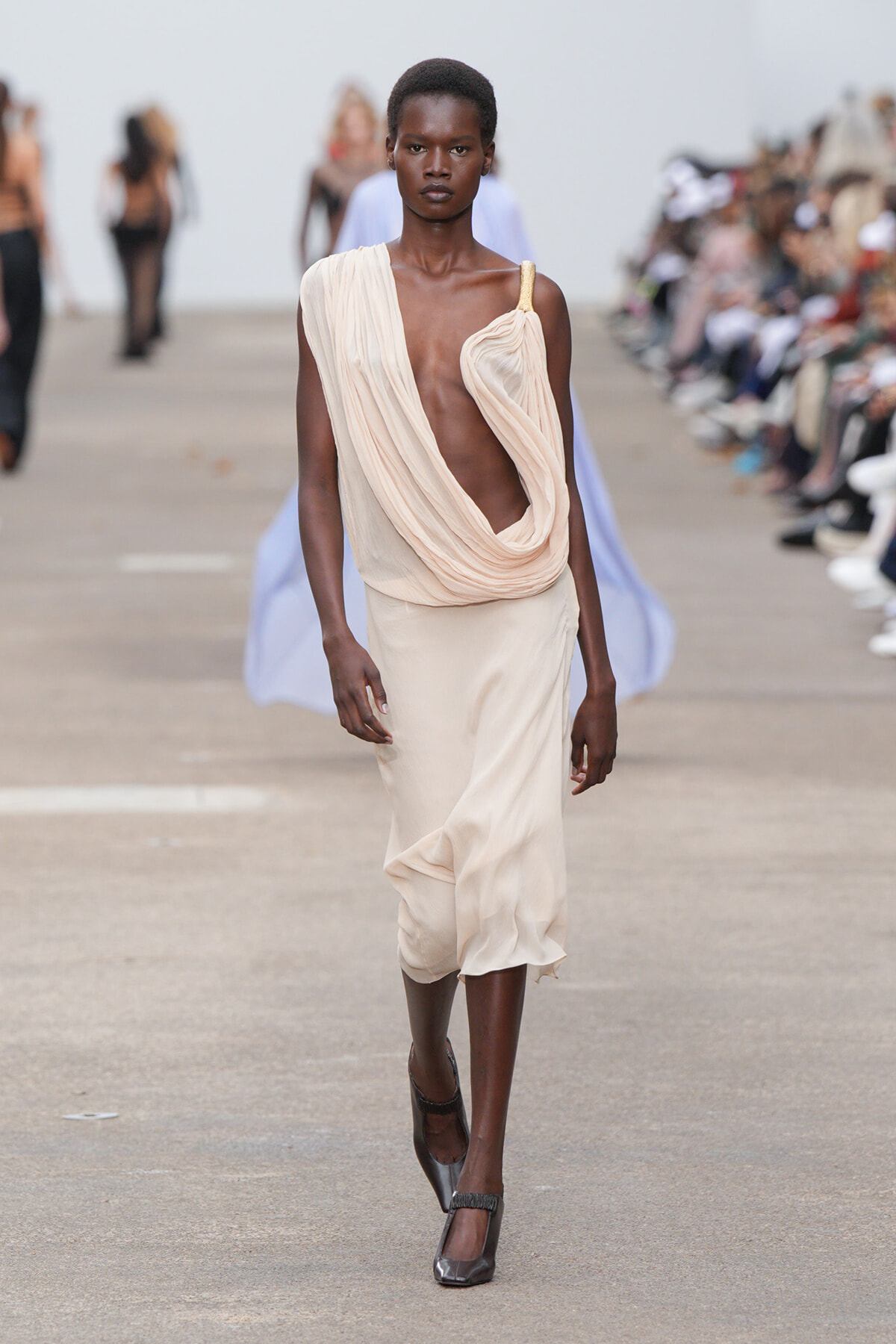 Model walking a runway in a sleeveless, draped beige dress with a gold strap and deep относительно neckline, wearing gray heels.