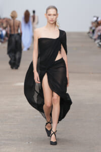 Model walking the runway in a black draped one-shoulder gown with a high side slit.
