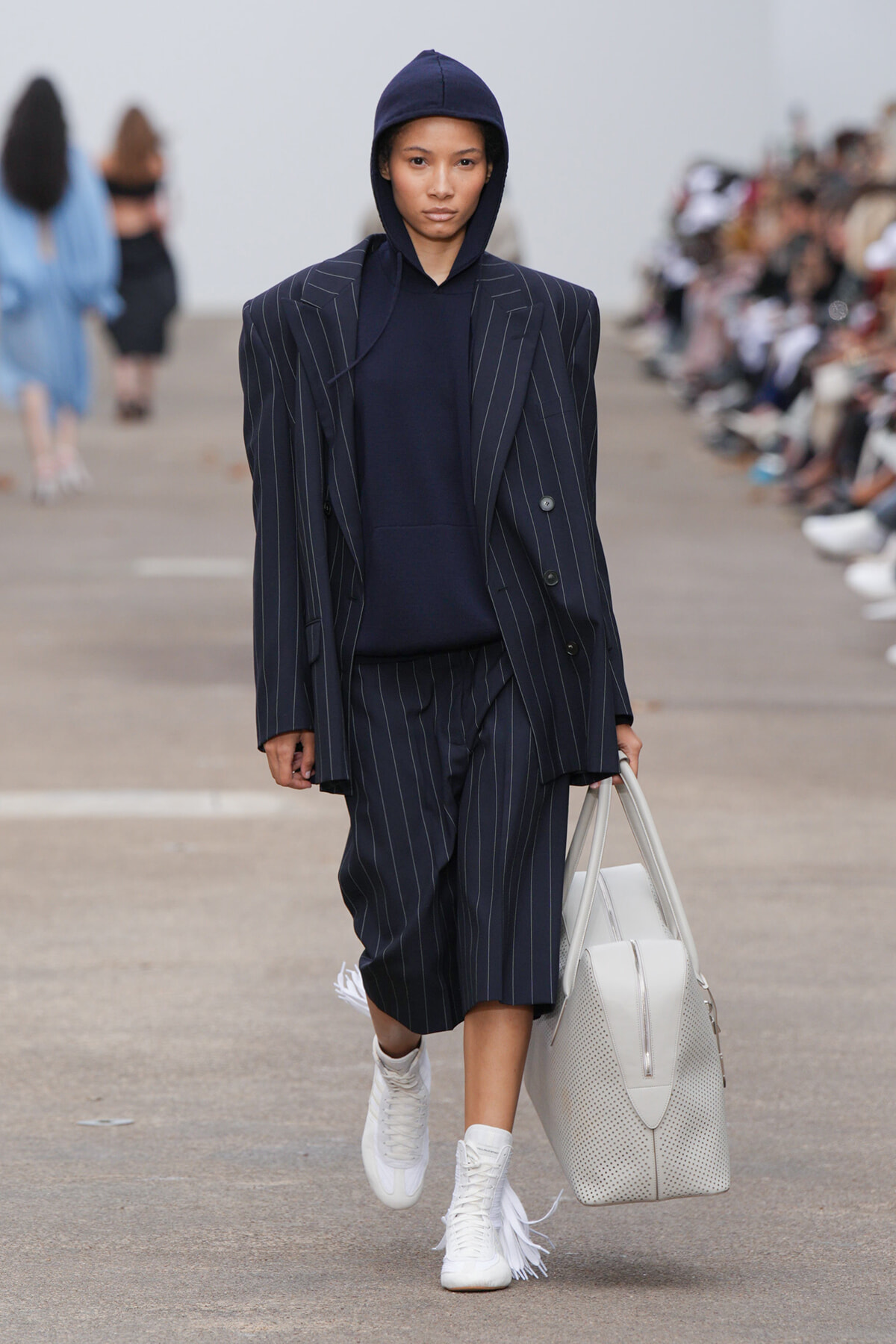 Model walking a fashion runway in a navy pinstripe oversized suit and matching navy hoodie, carrying a light gray perforated tote.