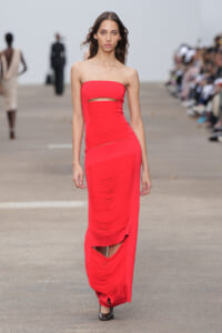 Model walks the runway in a bold red strapless gown with a waist cutout and ruched panels, floor-length and form-fitting.