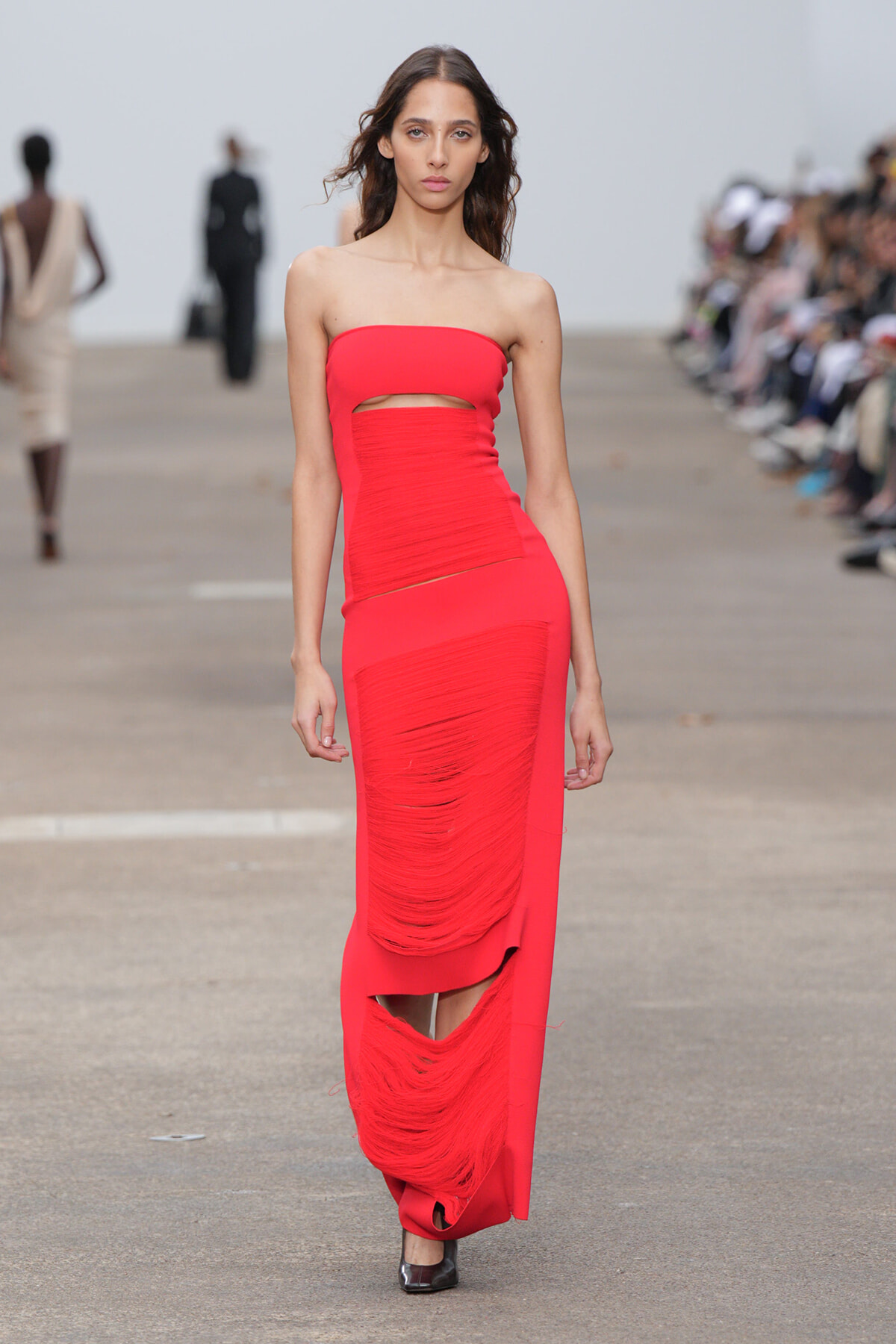 Model walks the runway in a bold red strapless gown with a waist cutout and ruched panels, floor-length and form-fitting.
