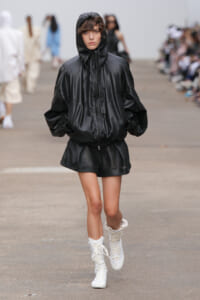 Model walking runway in an oversized black hooded jacket, black perforated shorts, and white lace-up boots.
