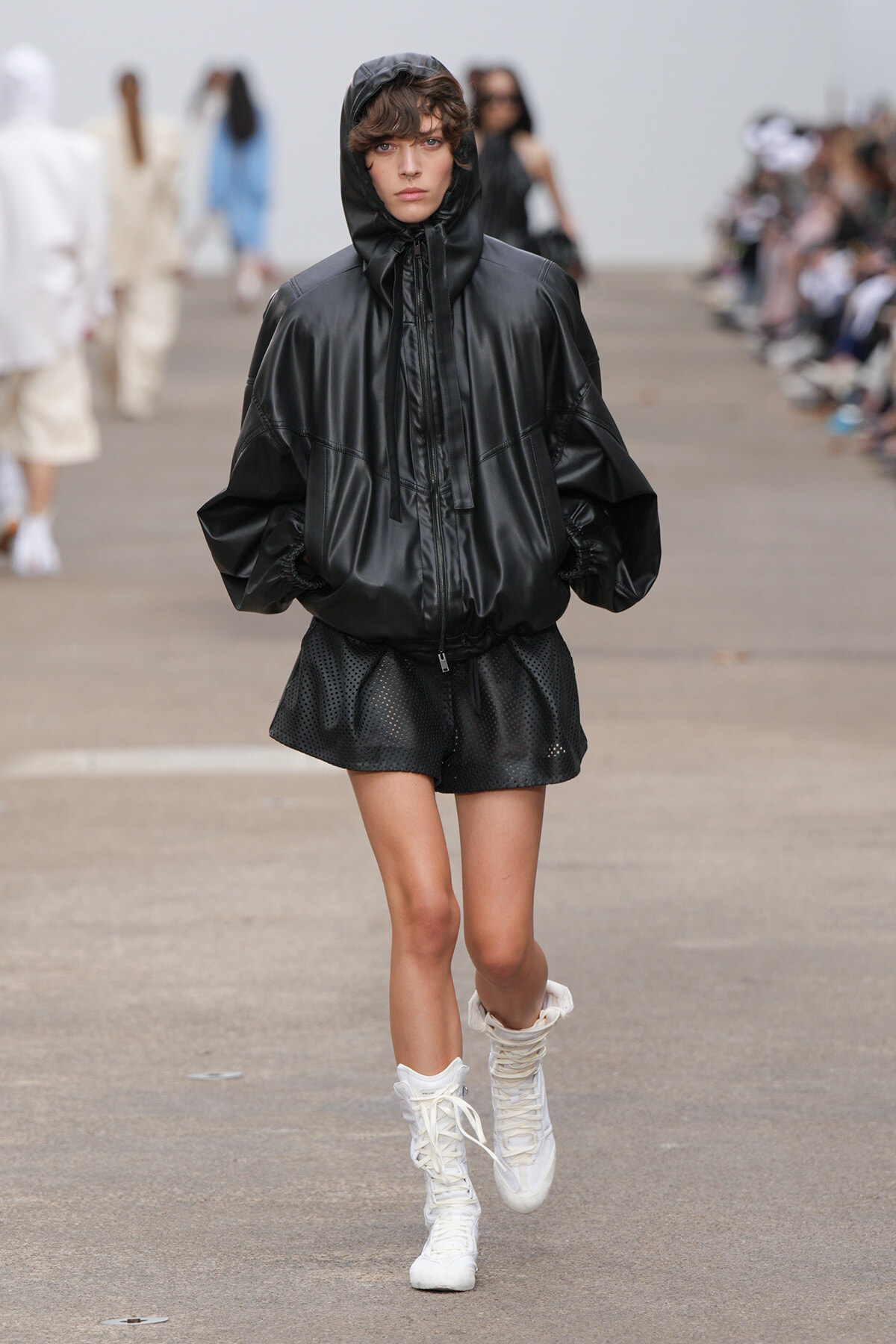 Model walking runway in an oversized black hooded jacket, black perforated shorts, and white lace-up boots.