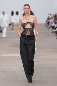 Model walks the runway in a black lace sheer bodysuit paired with high-waisted black pinstriped trousers.
