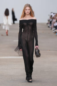Model walks the runway in a sheer black off-the-shoulder jumpsuit, carrying a small black handbag.