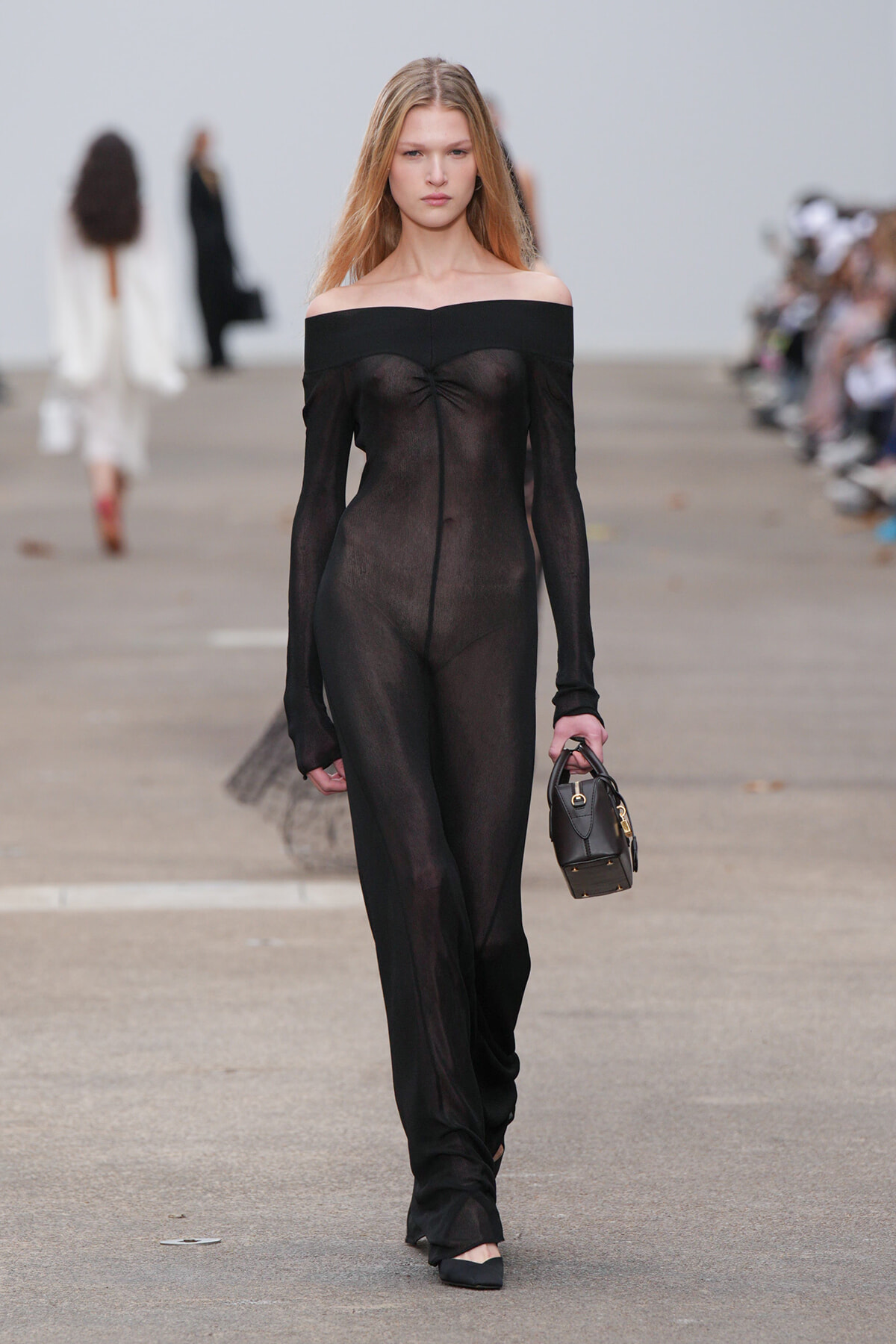 Model walks the runway in a sheer black off-the-shoulder jumpsuit, carrying a small black handbag.
