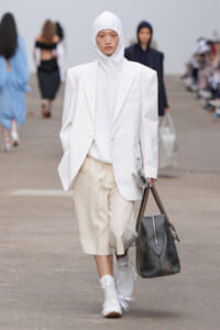 Model on a runway in an all-white outfit: oversized blazer and hoodie with beige shorts, white sneakers, and a large black bag.