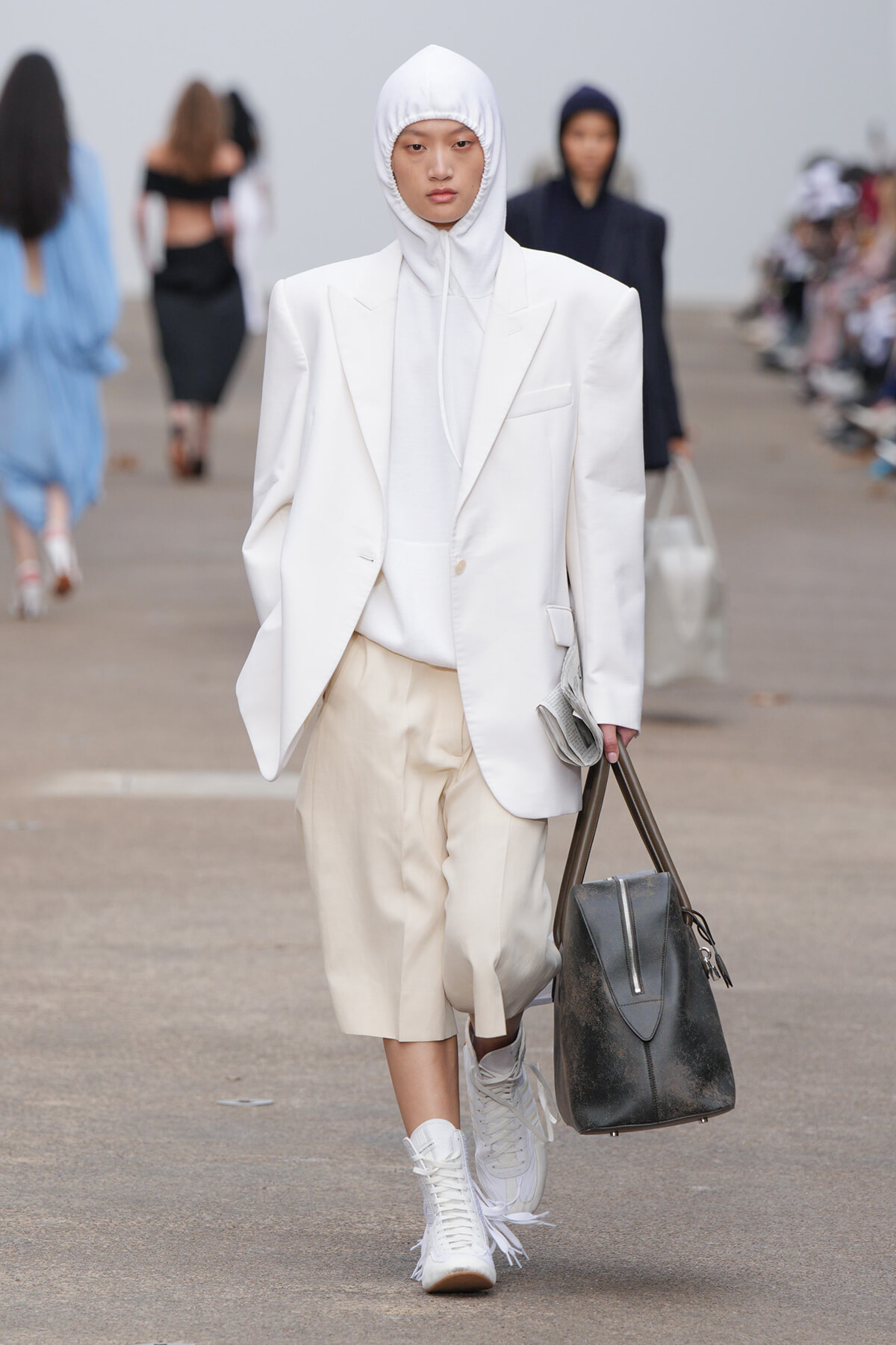 Model on a runway in an all-white outfit: oversized blazer and hoodie with beige shorts, white sneakers, and a large black bag.