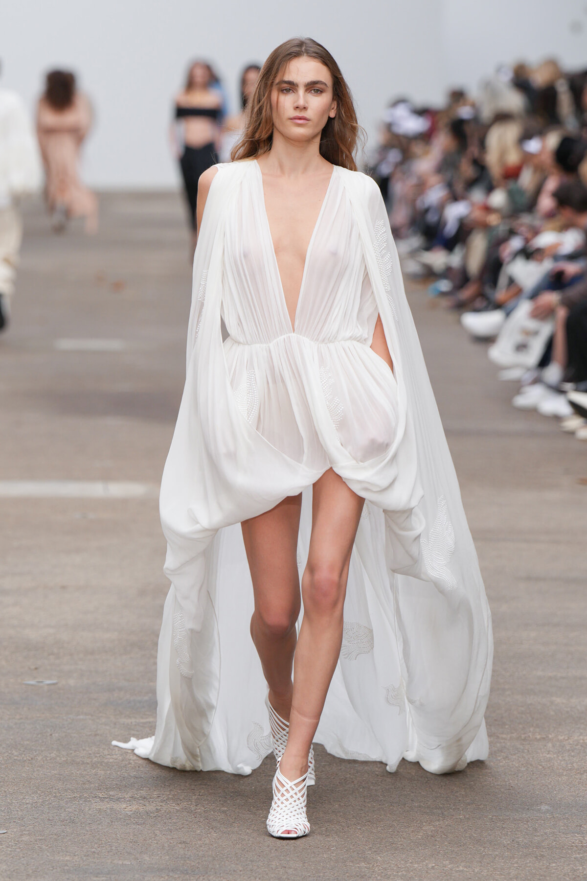 Model walking a runway in a white draped gown with a plunging V-neck and flowing cape sleeves.