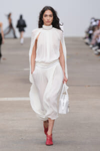 Model walking the runway in a sheer white pleated gown with cape sleeves, carrying a white handbag and wearing red lattice heels.
