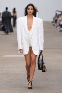 Runway model in an oversized white blazer with a deep plunge, carrying a black handbag and wearing black strappy heels.