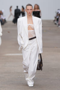 Model walking a runway in an oversized white suit with a sheer lace bralette, belt, and white trousers, carrying a dark quilted handbag as she strides forward.