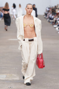 Model on a fashion runway wearing a loose beige suit with a gold bralette and a bright red handbag.