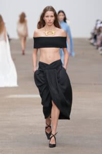 Model walking a runway in a black off-shoulder bandeau top with a draped black skirt and a gold statement necklace on heels.