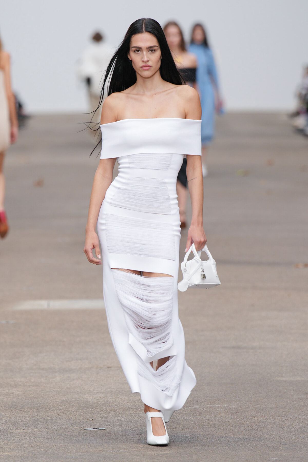Female model walks a fashion runway in a white off-the-shoulder dress with shredded panels, carrying a small white handbag.