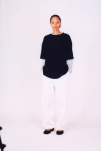 Person standing against a white background wearing a black short-sleeve top over light sleeves, white pants, and black flats.