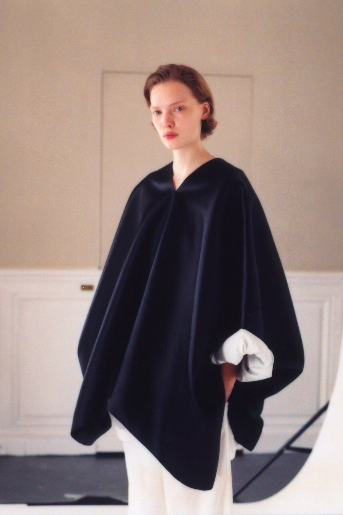 Model wearing a voluminous black cape over a white-sleeved shirt, standing indoors in a neutral room.