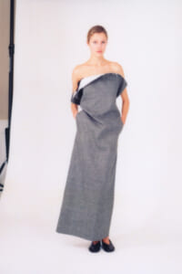 Model posing in a strapless, gray textured dress with a white underlayer, hands in pockets, against a white studio backdrop.