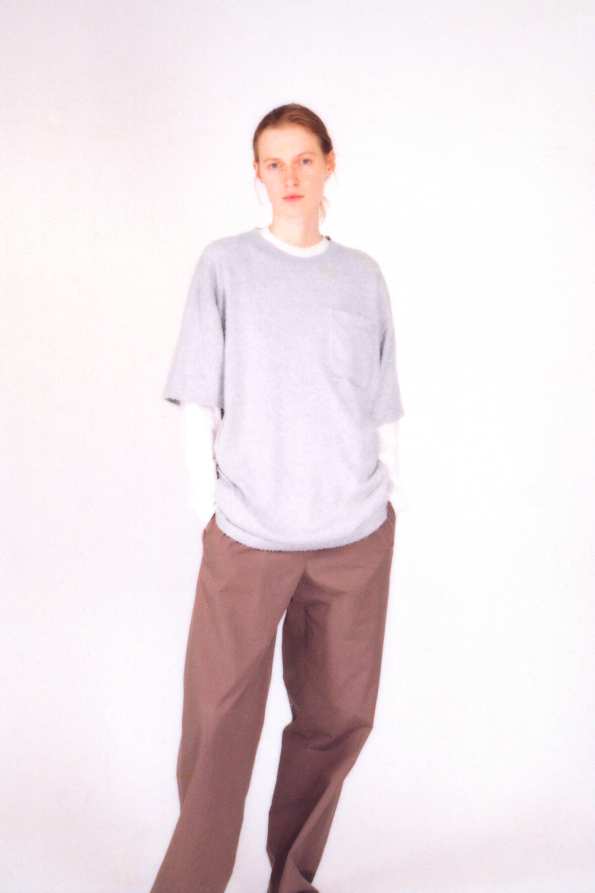 Person standing in a light gray oversized T-shirt over a white long-sleeve shirt, brown wide-leg pants, neutral studio background.