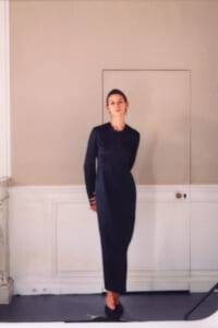 Woman in a navy long-sleeve sheath dress stands with hands behind her back in a plain, light-colored room.