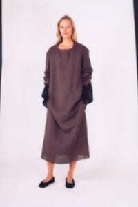 Person standing in a loose brown, ankle-length dress with black gloves and black flats, against a white backdrop, neutral expression.