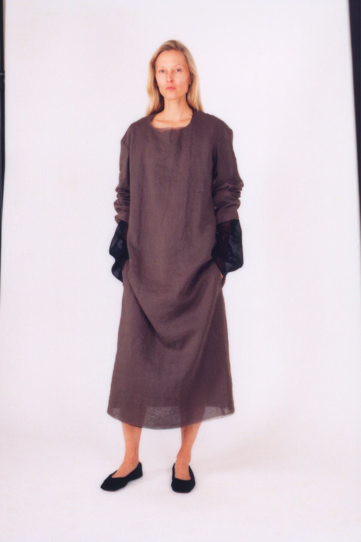 Person standing in a loose brown, ankle-length dress with black gloves and black flats, against a white backdrop, neutral expression.
