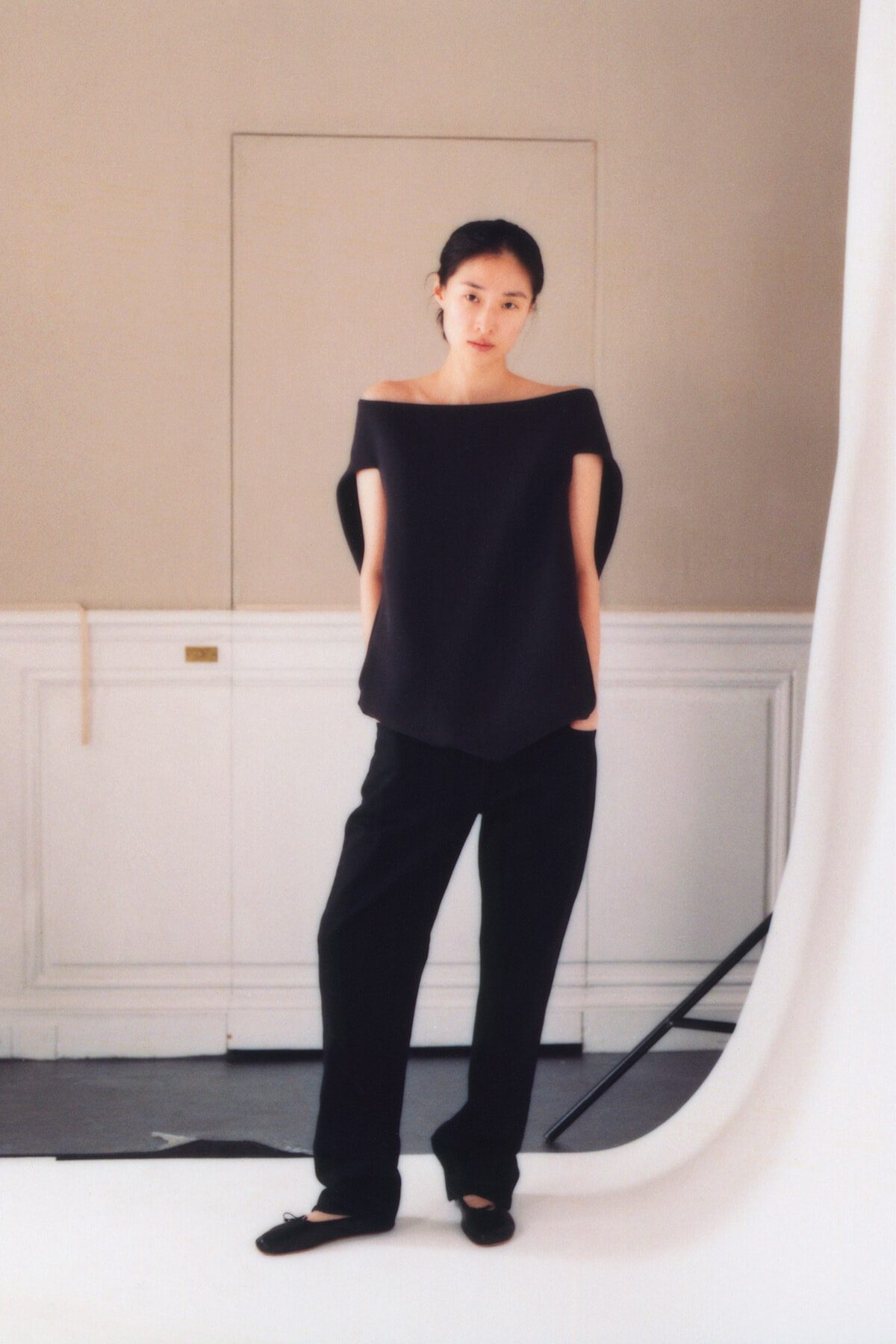 Full-length portrait of a woman in a black off-shoulder top and black pants standing in a studio with a white backdrop.