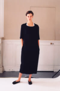 Model wearing a black midi dress with a white collar, standing in a studio with hands in pockets.