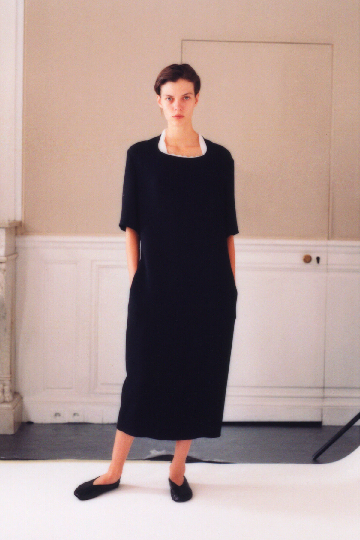 Model wearing a black midi dress with a white collar, standing in a studio with hands in pockets.