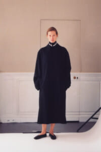 Person wearing a long black coat with a high collar, standing in a minimalist studio setting with hands in pockets.