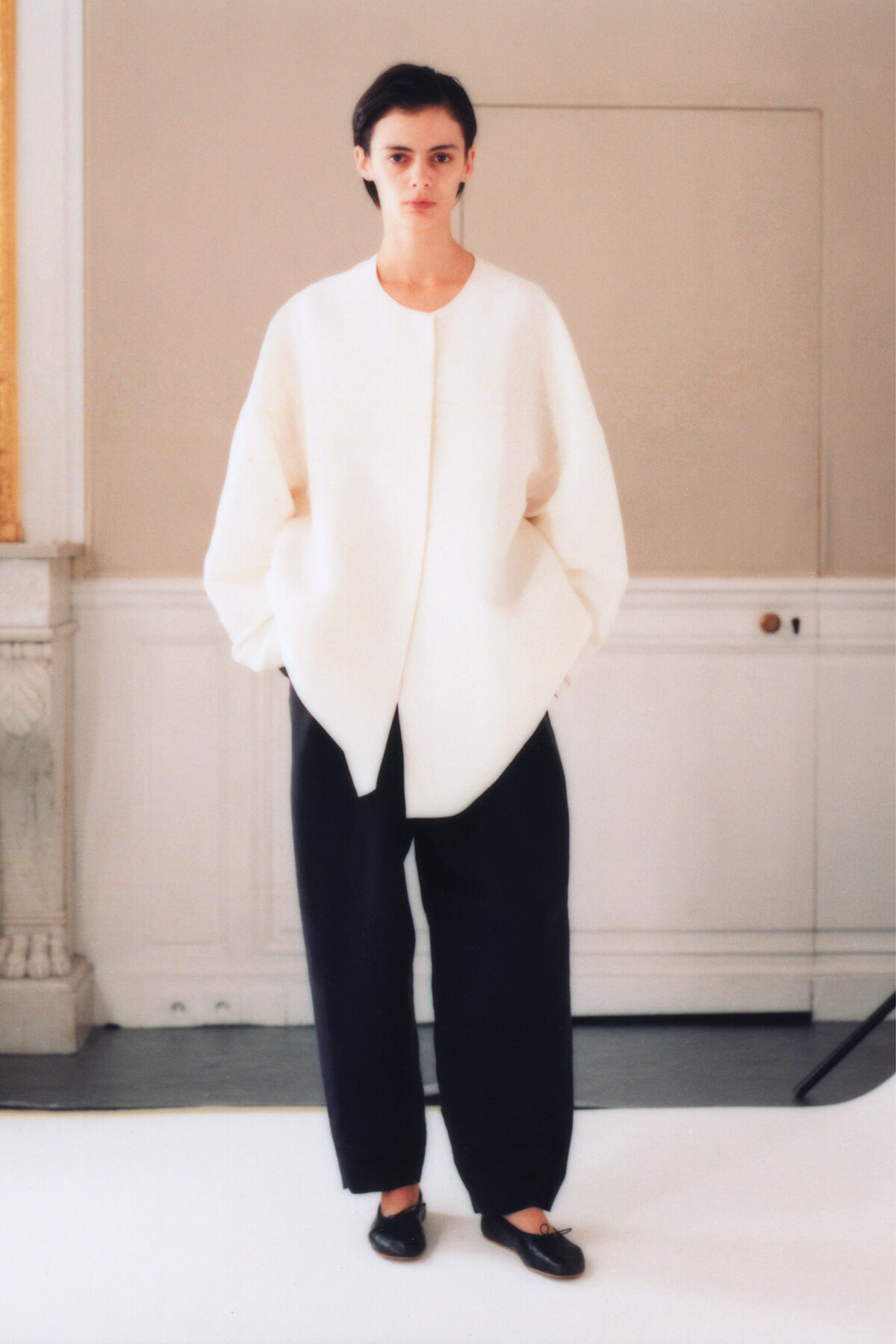 Model stands in a studio wearing an oversized white coat-like top and black wide-leg pants, with hands in pockets.