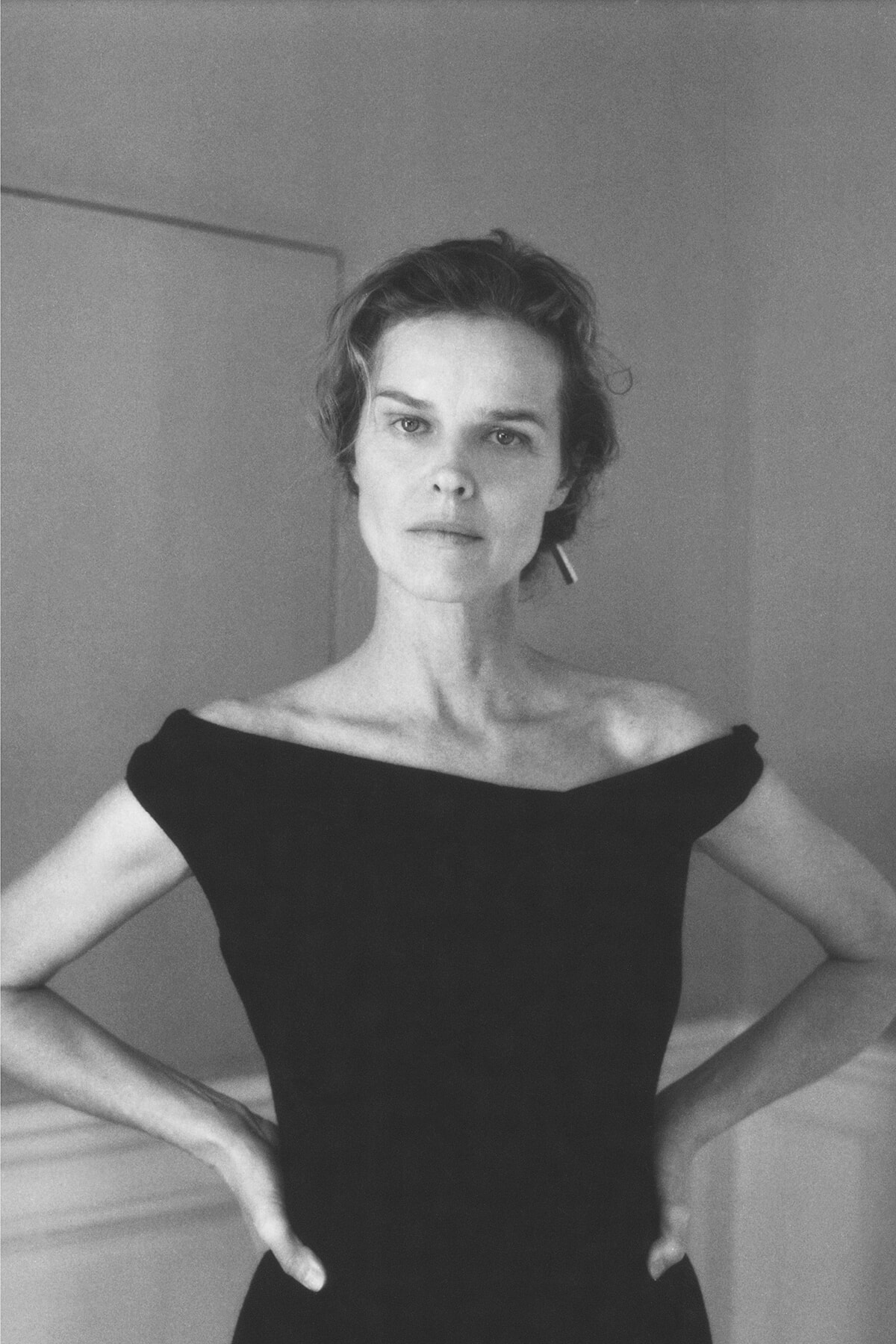 Black-and-white portrait of a woman with short hair wearing an off-the-shoulder black dress, standing with hands on hips and a serious expression.