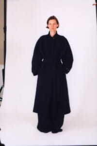 Woman standing in a long black belted coat with hands in pockets against a white studio backdrop
