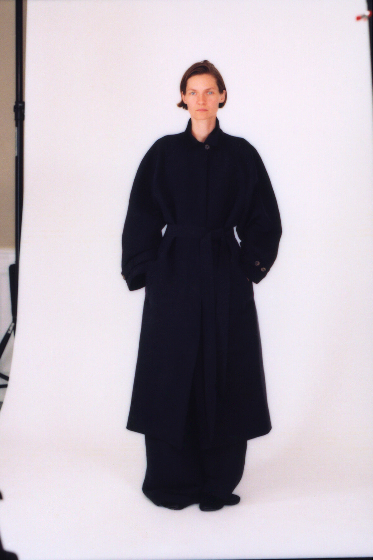 Woman standing in a long black belted coat with hands in pockets against a white studio backdrop
