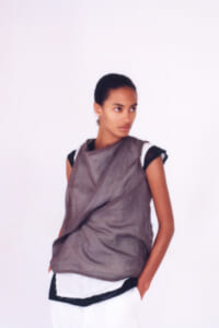Model wearing a loose taupe draped top over black-and-white layered clothing, standing with hands in pockets against a white backdrop.